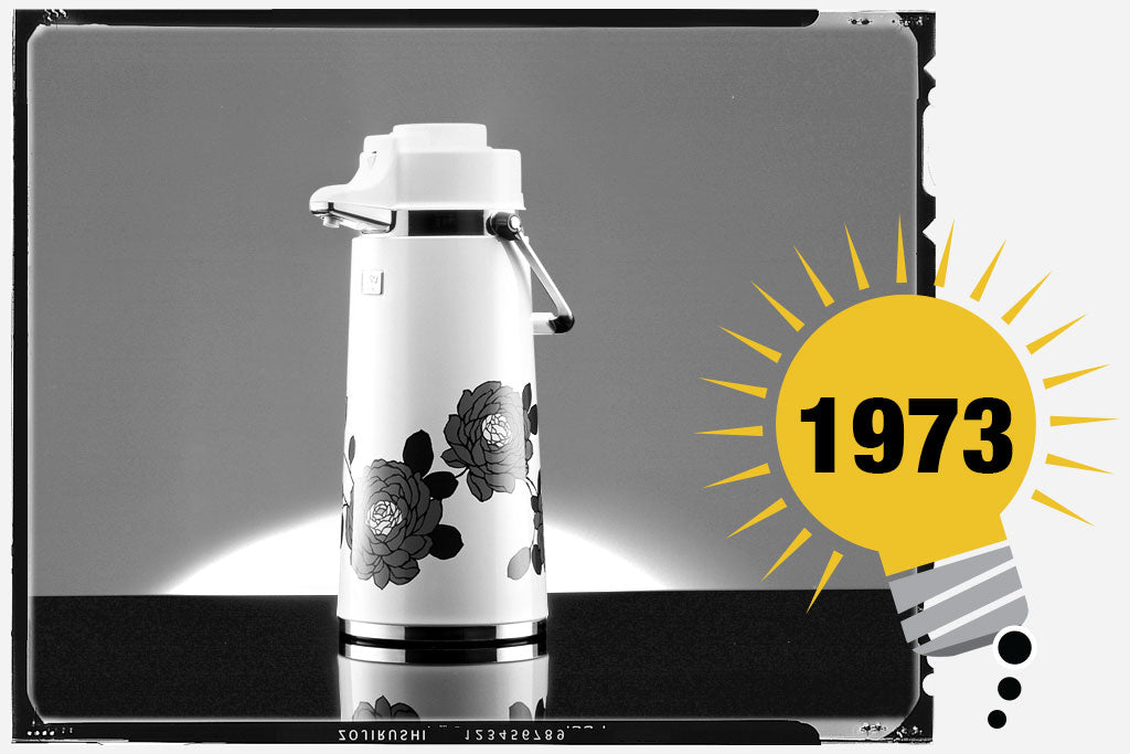 Black and white photo of the Zojirushi Air Pot® Dispenser. The unit is white with a large floral design around the body. Photo is labeled with the year 1973.