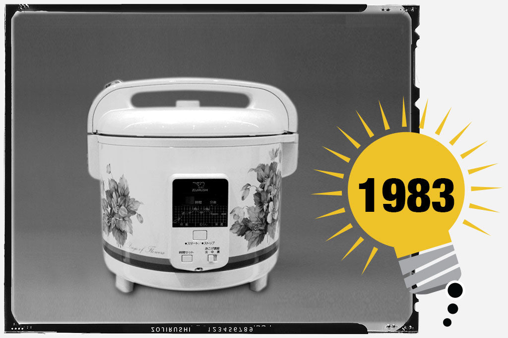 Black and white photo of the first Zojirushi Micom Rice Cooker & Warmer. The unit is white with floral prints around it’s body, and has a square panel with switch in the front. Photo is labeled with the year 1983.