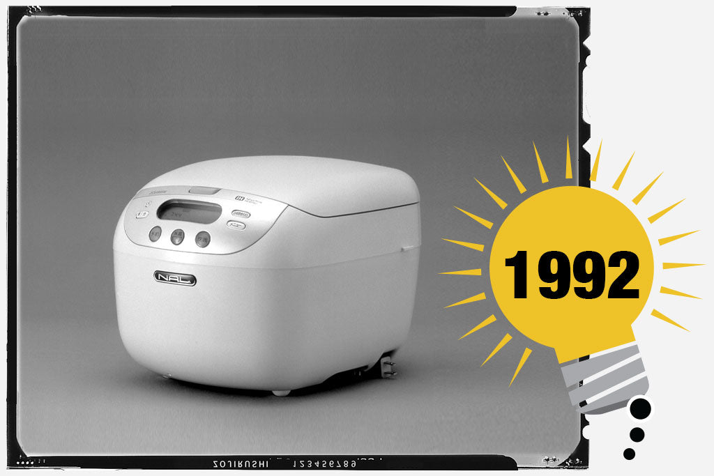 Black and white photo of the first Zojirushi Induction Heating Rice Cooker & Warmer. The unit is white and cube-like shape, with a shiny control panel. Photo is labeled with the  year 1992.