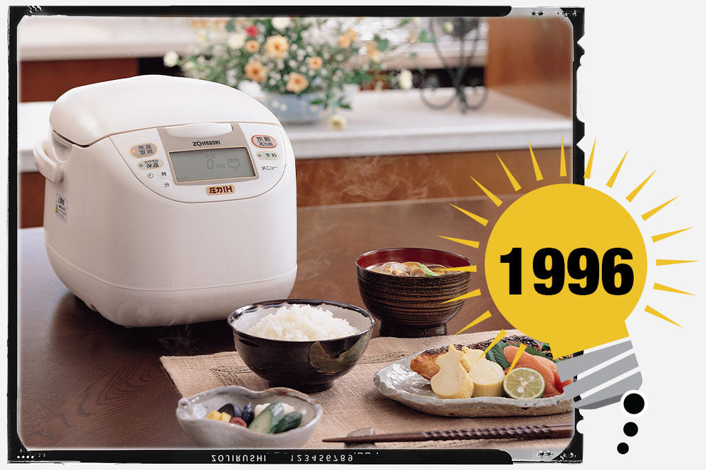First Zojirushi Pressure IH Rice Cooker & Wamer that is placed on a table next to a bowl of rice, which is a part of the Japanese meal. Photo is labeled with the year 1996.