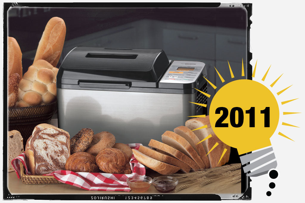 Zojirushi Home Bakery Virtuoso® Breadmaker with various type of bread around the unit. The unit is stainless and black. Photo is labeled with the year 2011.